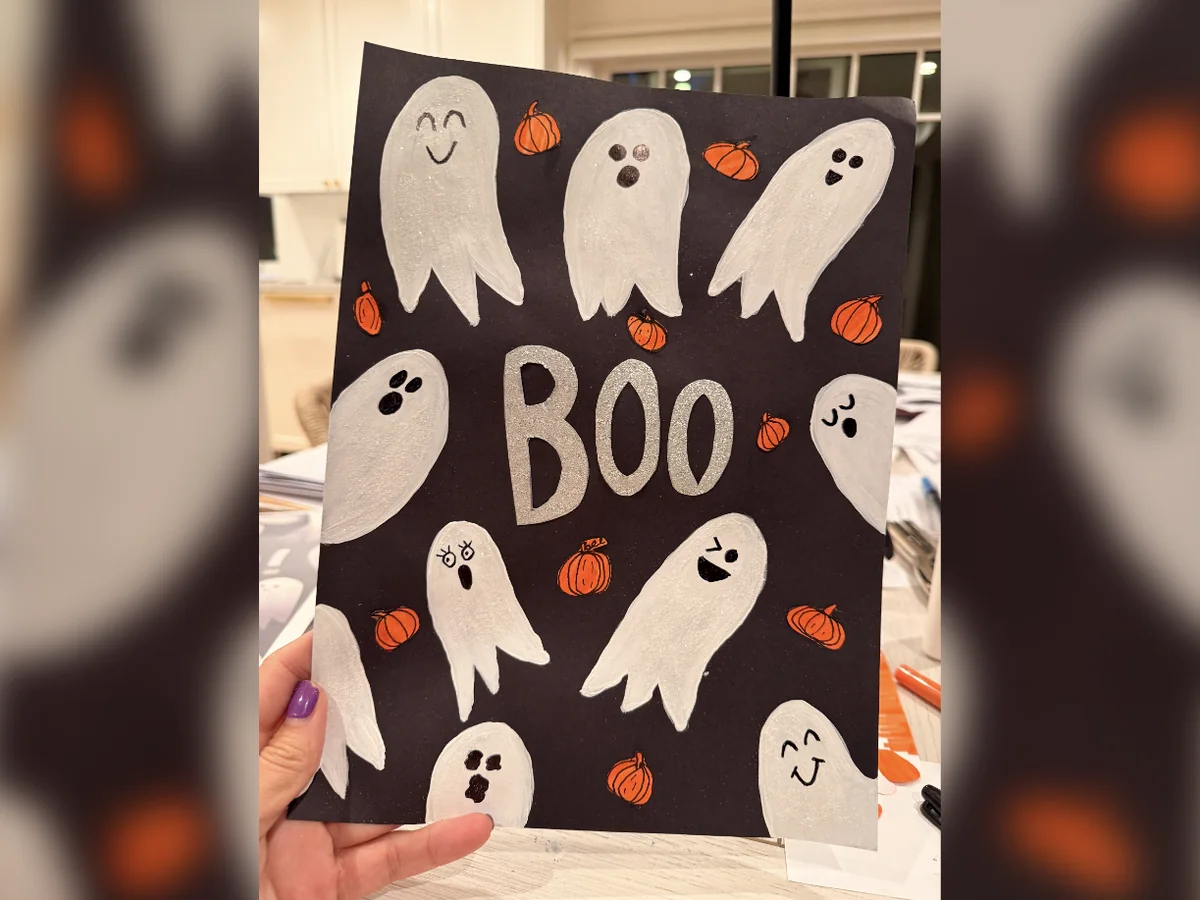 The finished October art project: white glitter ghosts with drawn faces and little orange pumpkins around the word BOO on black paper