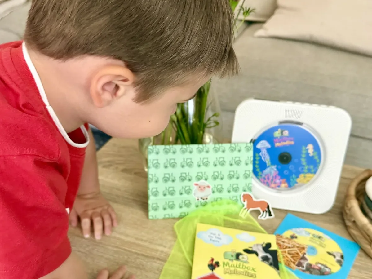 A young child leaning in to listen to the Mailbox Melodies CD on a portable player, with the booklet and stickers laid out beside it